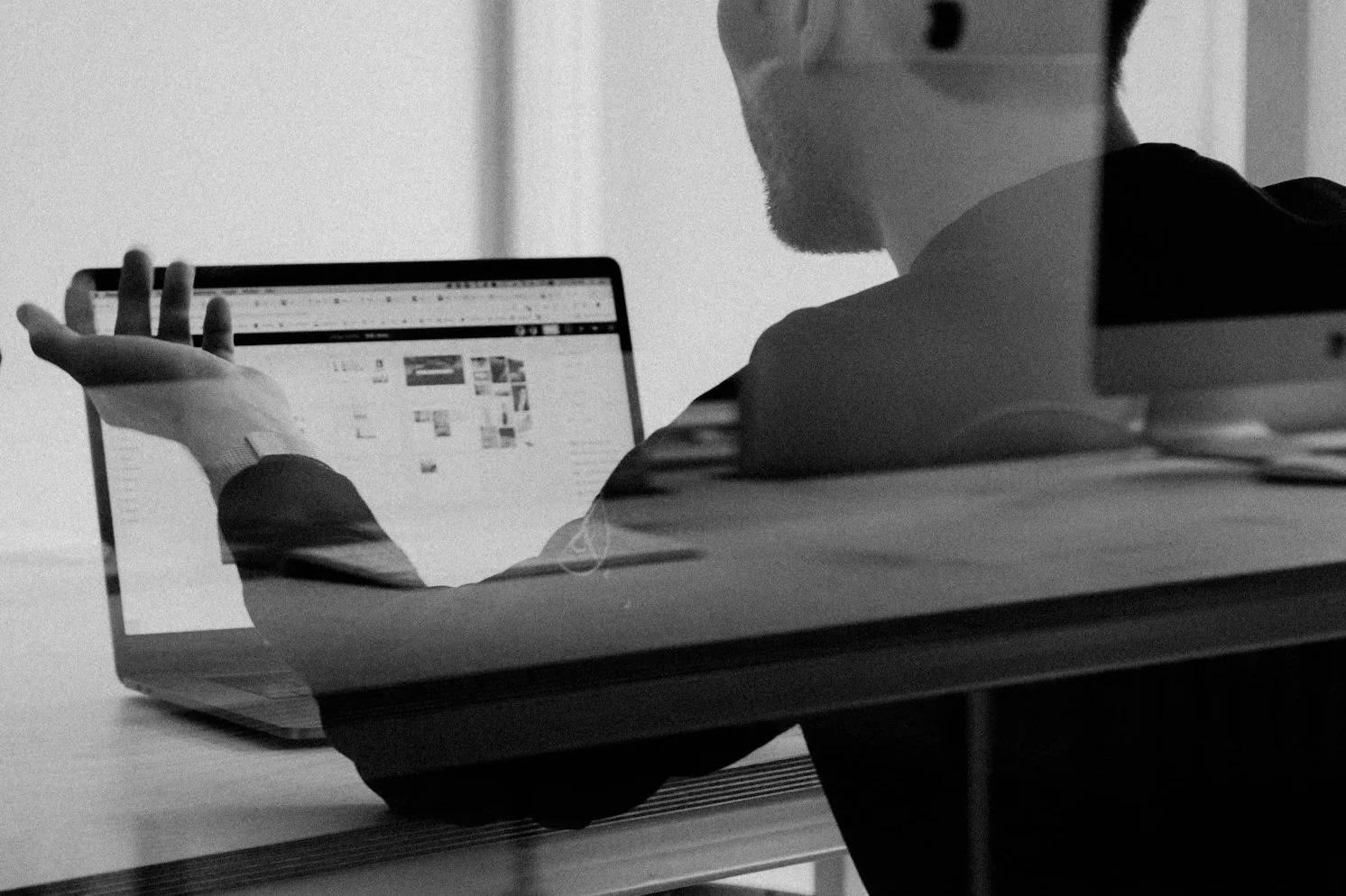 A person in a black jacket sitting at a desk, working on a laptop, seen through a glass reflection, in black and white.