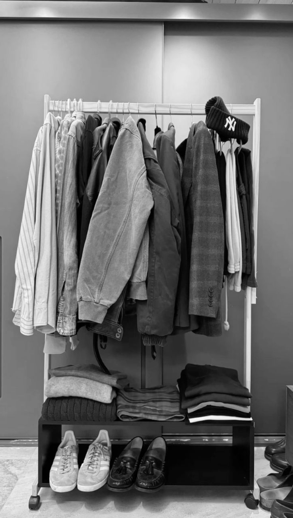 A clothing rack with shirts, jackets, and sweaters, topped with a knit winter hat, and a shelf with folded clothes and shoes underneath.