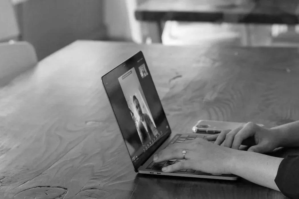 Person using a laptop and smartphone on a wooden table in a home or office setting.