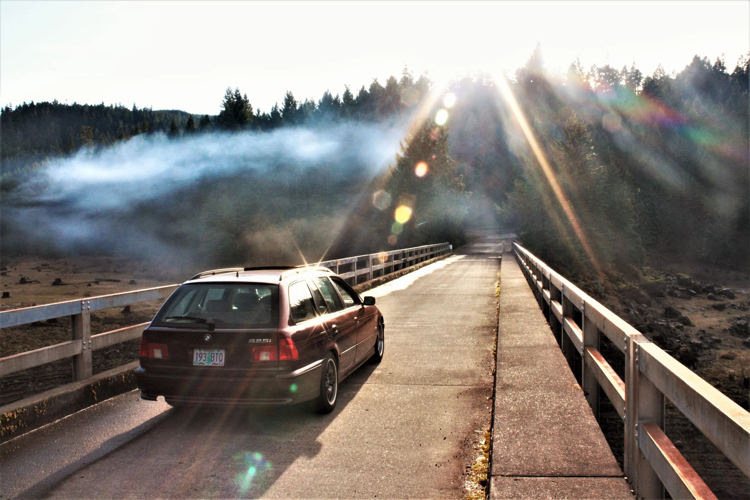 A red station wagon parked on a narrow bridge over a river or gorge, with trees and hills in the background, and sunlight creating lens flares and reflections.
