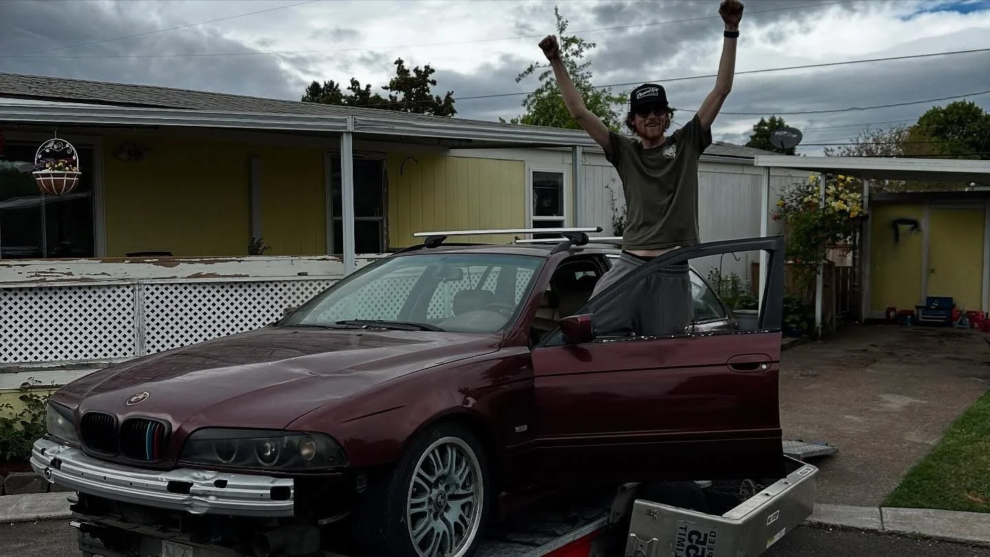 A man with long hair and glasses, wearing a black cap, stands on a car with his arms raised in celebration. The car is a maroon BMW with some front bumper damage, parked on a trailer in a residential driveway. The background shows a house, some trees, and a cloudy sky.