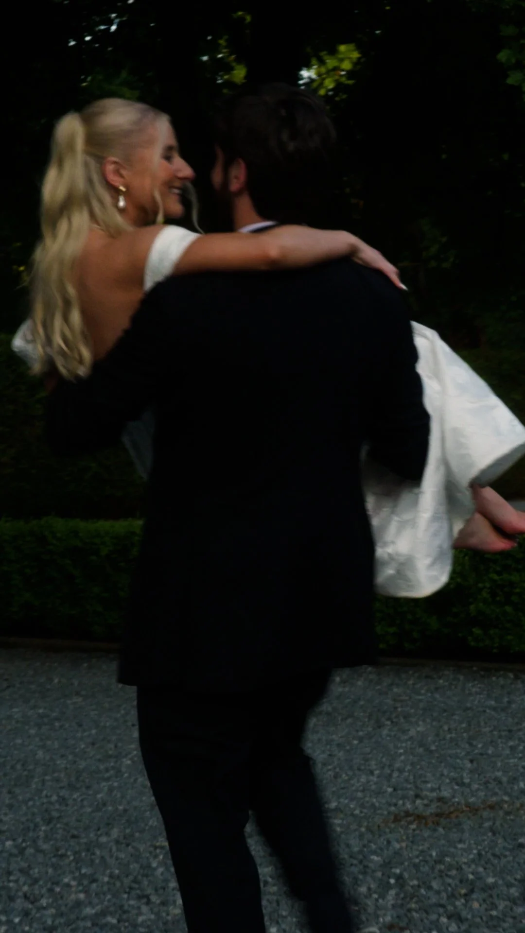 A man in a black suit carrying a woman in a white dress, dancing outdoors at night.