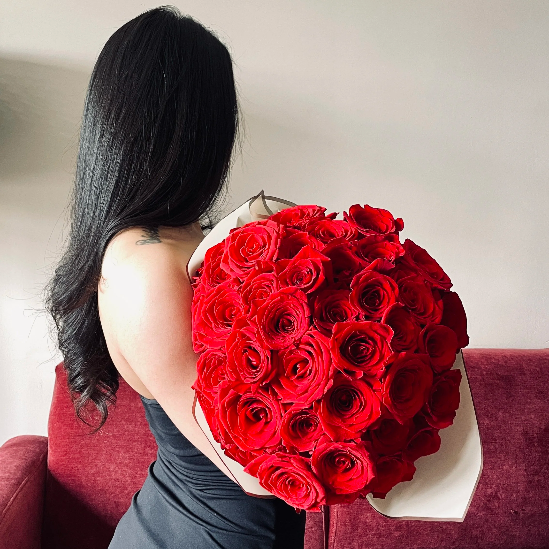 Woman with long black hair holding a large bouquet of red roses, sitting on a red couch, facing away from the camera.