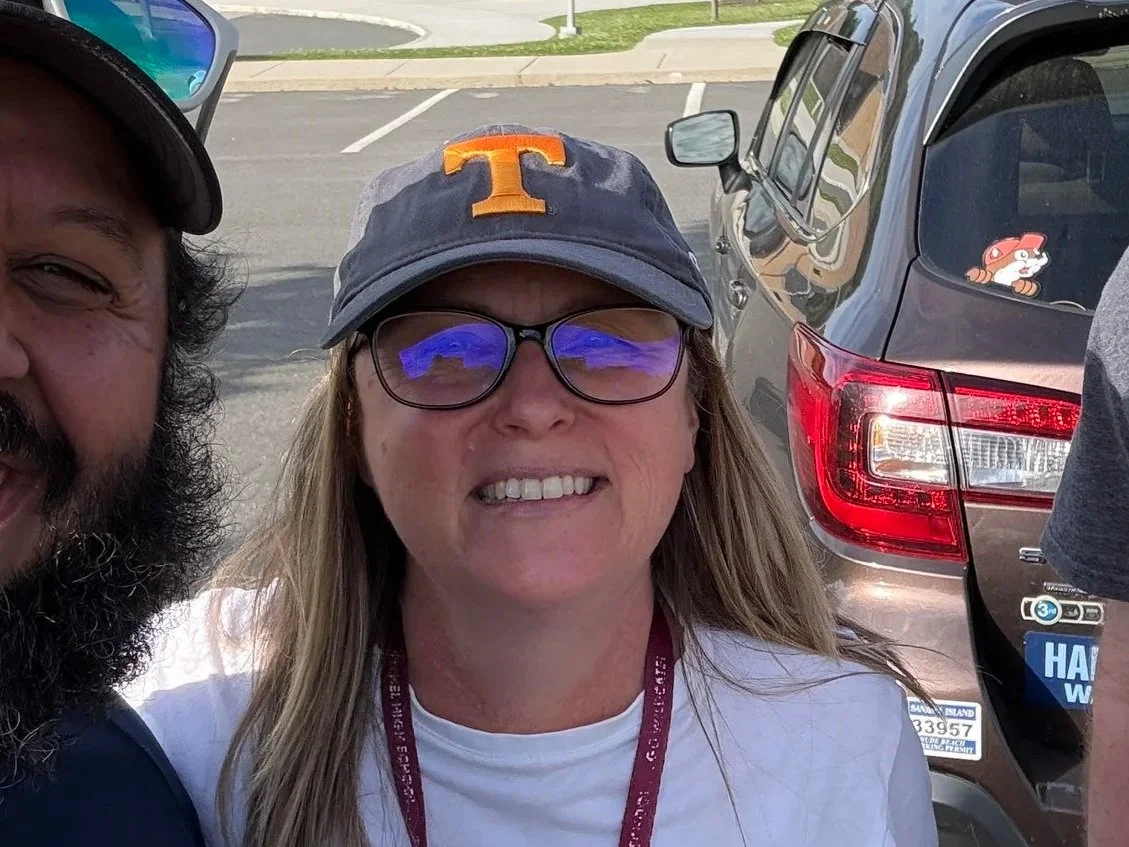 A woman wearing glasses, a baseball cap with an orange 'T', and a white t-shirt, smiling for a selfie. Part of a man with a beard and another person are also visible. They are outdoors in a parking lot with cars in the background, one with a sticker of a cartoon dog in the rear window.