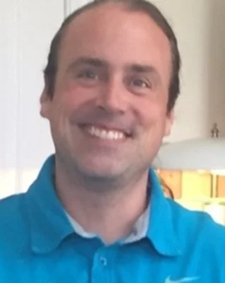 A smiling man wearing a blue polo shirt, standing indoors with part of a kitchen visible in the background.
