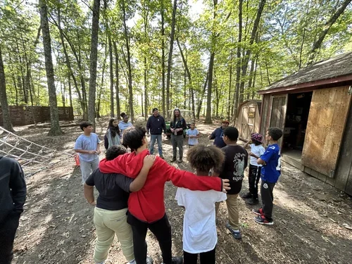 Group of children and adults gathered outdoors in a wooded area, some hugging, near a shed.