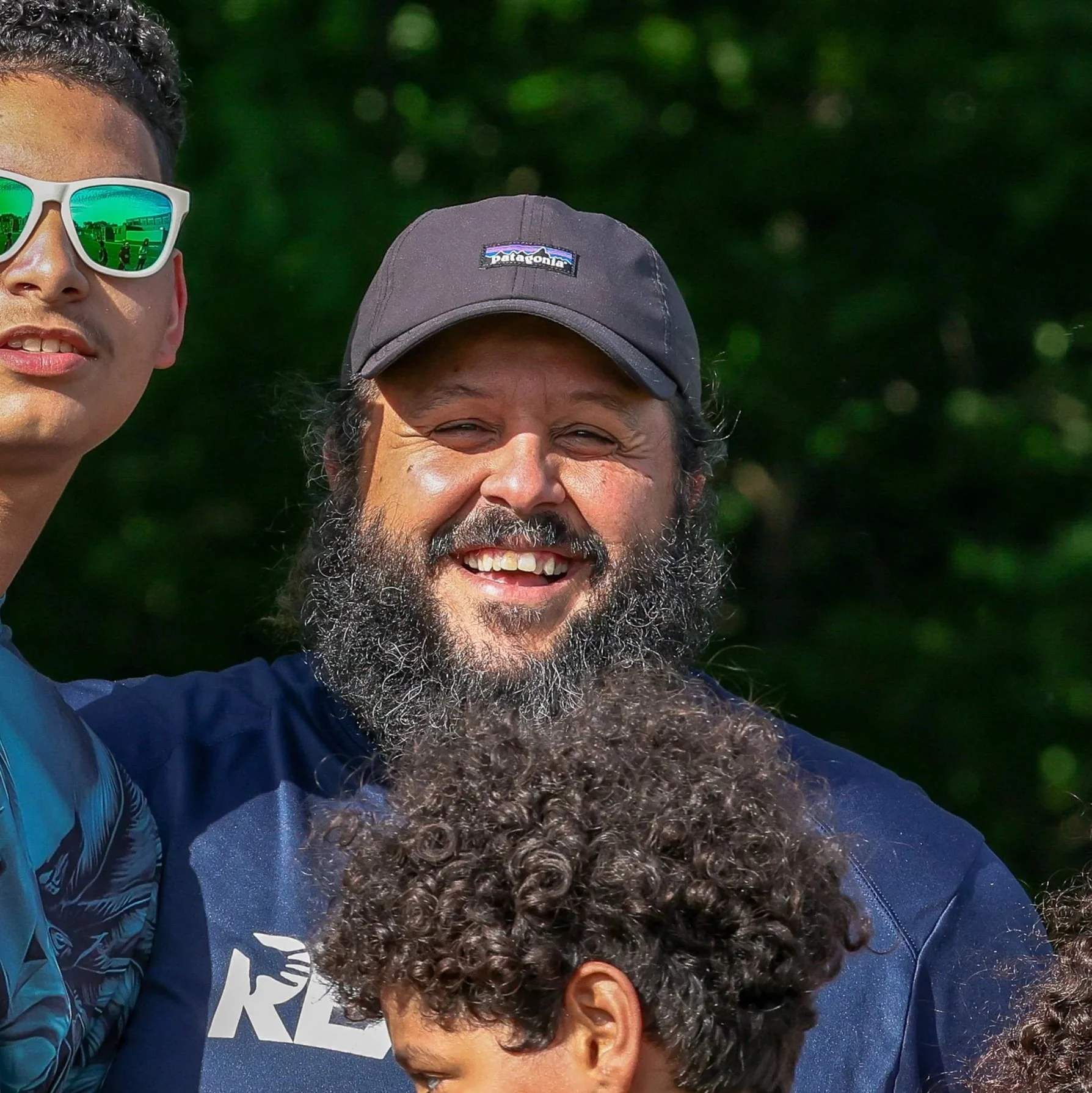 Close-up photo of two smiling men outdoors with green foliage in the background. The man on the left wearing white sunglasses and the man on the right with a gray Patagonian cap and a full beard.