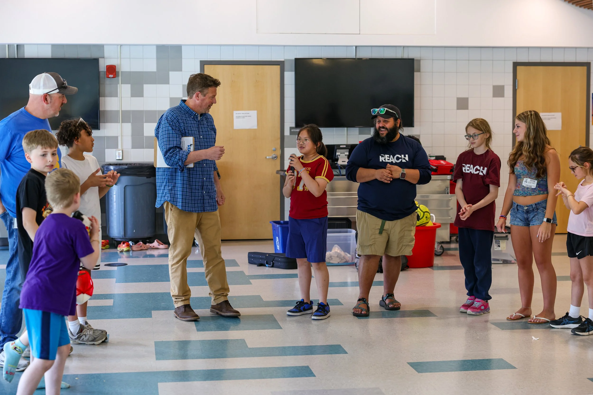 Group of children and adults gathered indoors in a school hallway, with some holding toys or balls, engaged in conversation and smiling.