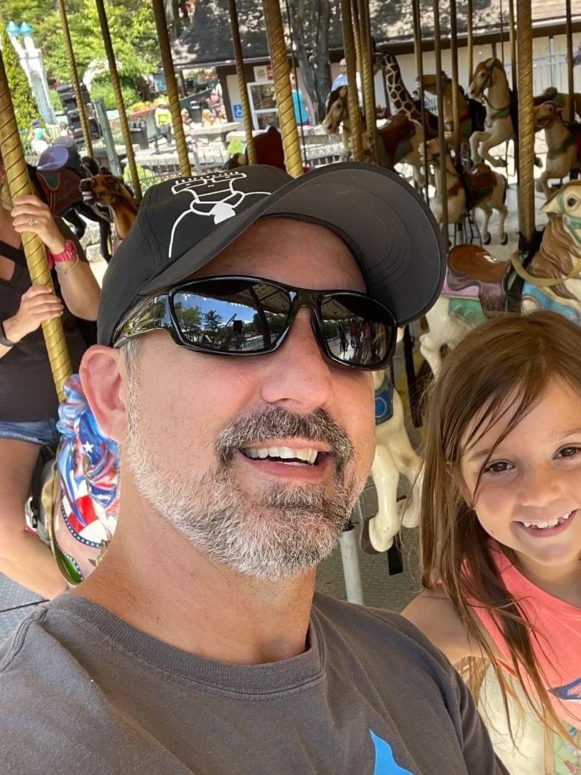 A man wearing a black cap with a white design, black sunglasses, and a gray T-shirt, smiling and taking a selfie on a carousel ride. A young girl with brown hair and a pink T-shirt is smiling next to him. Behind them, there are carousel horses and an outdoor amusement park setting.