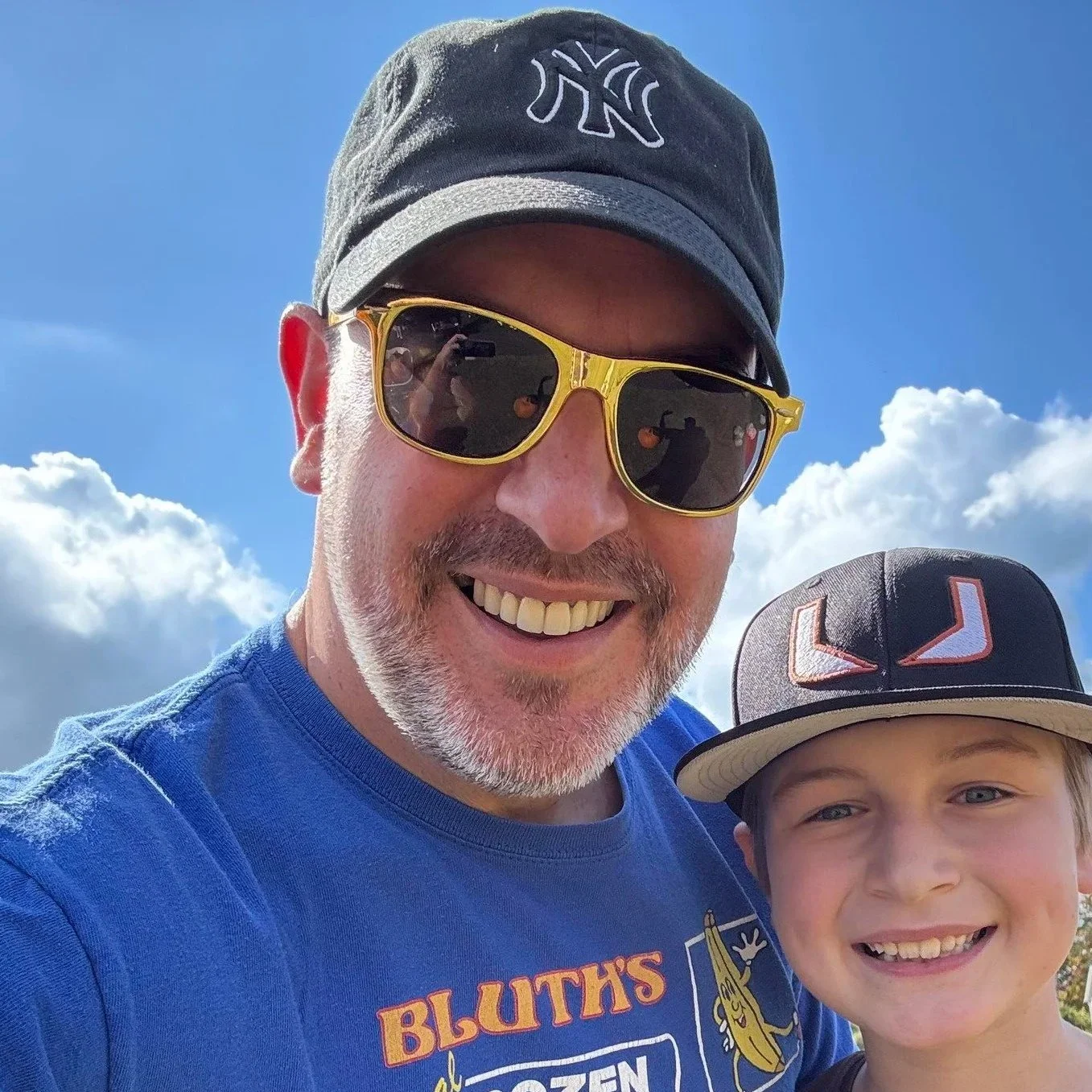 A man with glasses and a cap smiling next to a young boy wearing a cap, outdoors with a blue sky and clouds in the background.