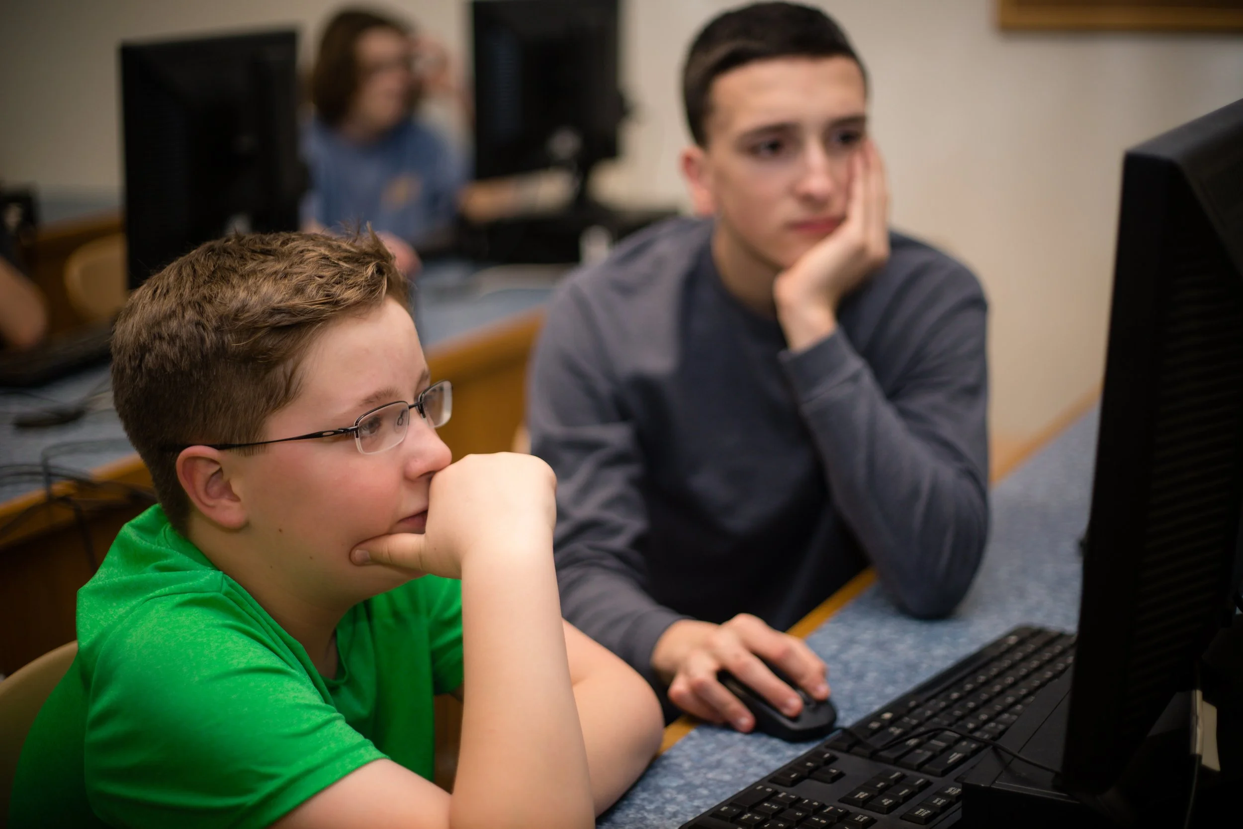 Two boys are working on computers in a classroom. One is wearing glasses and a green shirt, focused on the screen. The other, with dark hair, is resting his chin on his hand, looking at the monitor. In the background, a woman with glasses is also working on a computer.