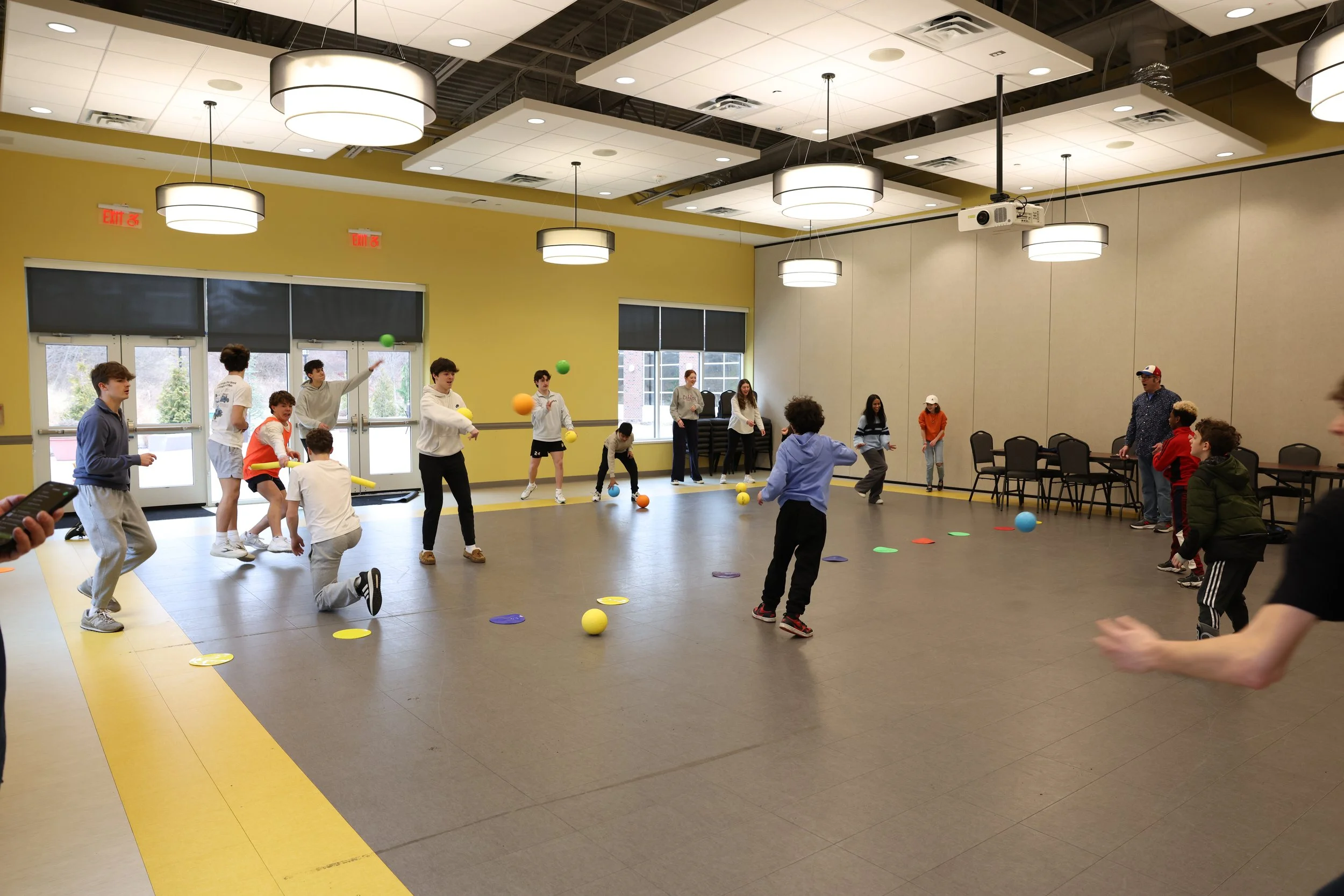 Children and teenagers playing a game indoors, juggling colorful balls, with some standing and some kneeling, in a large room with yellow walls, large windows, and hangings lights on the ceiling.