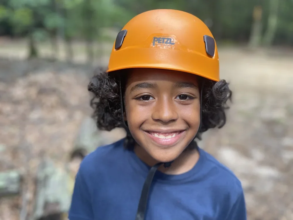 Smiling boy in a blue shirt wearing an orange climbing helmet outdoors in a wooded area.