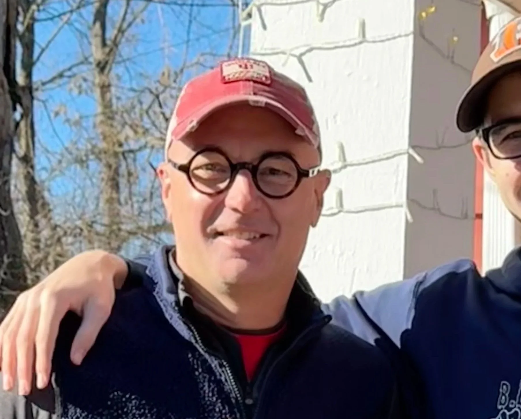 Two men happy, wearing glasses and baseball caps, with a tree and blue sky behind them, outdoors during daytime.