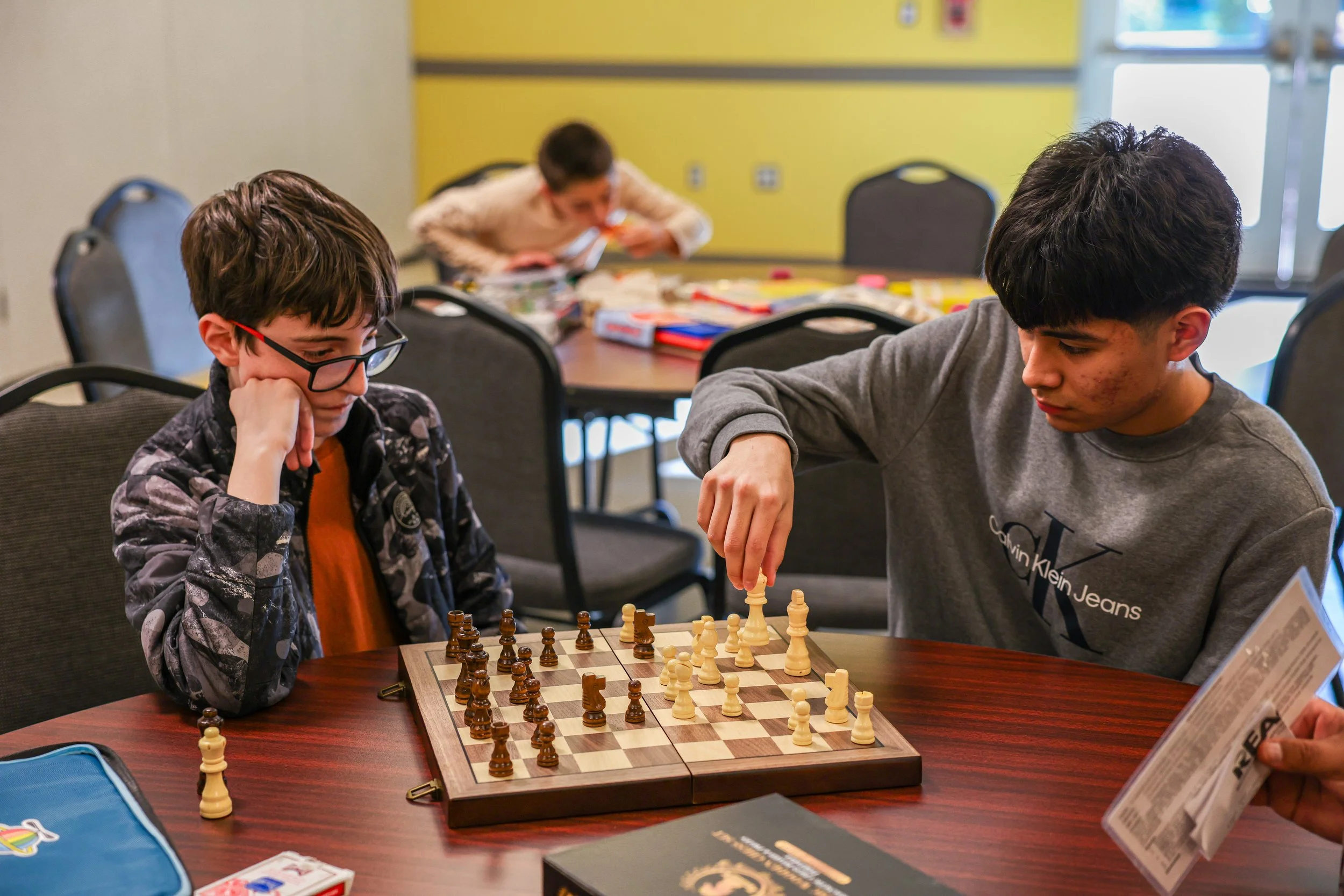 Two boys playing chess at a table, one wearing glasses and an orange shirt under a patterned jacket, the other with dark hair and a gray Calvin Klein hoodie.