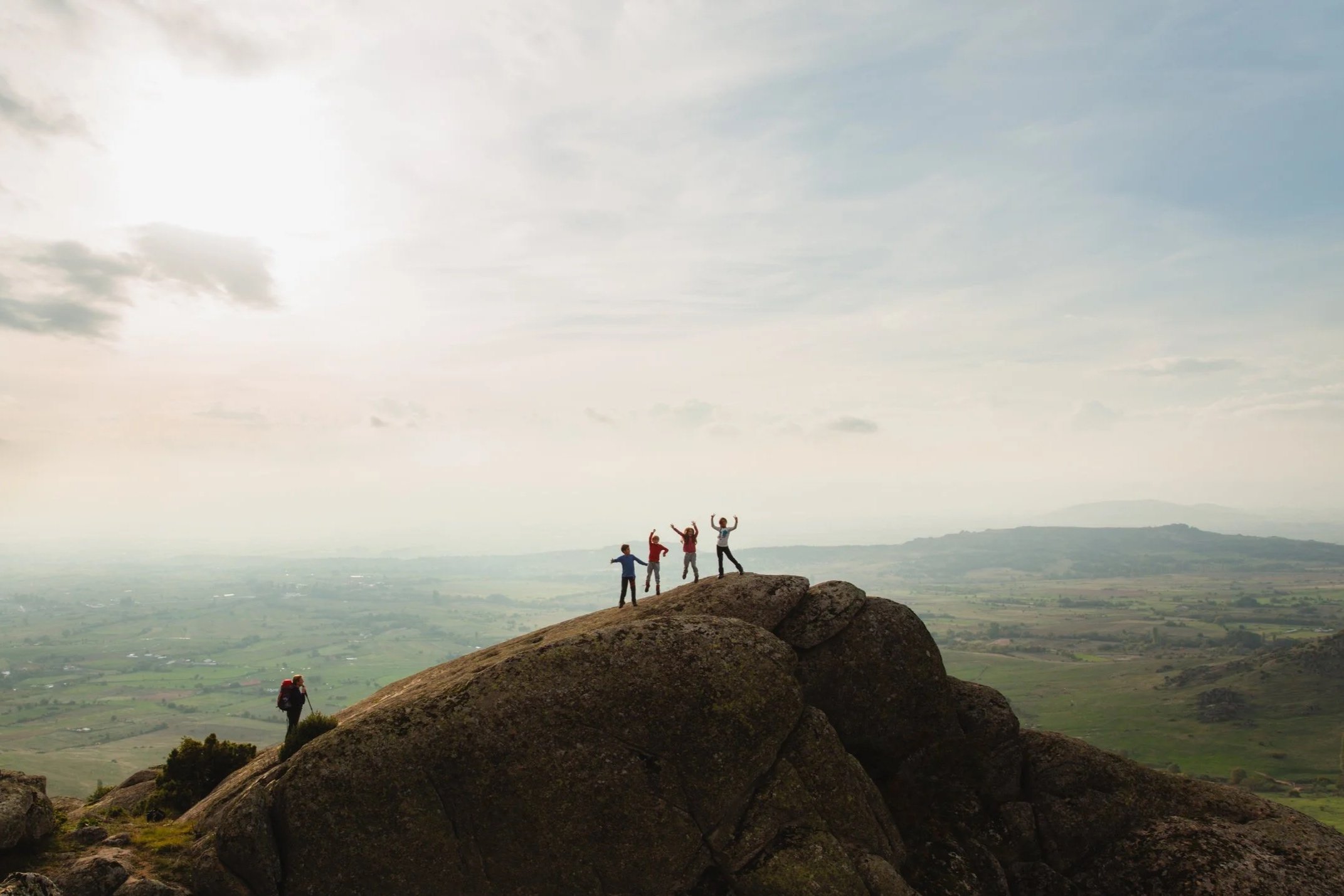 Four children standing on a large rock formation, with one child leading, in an open landscape with hills, and an adult hiker at the base of the rocks, while a few others are further back, enjoying a scenic outlook on a cloudy day.