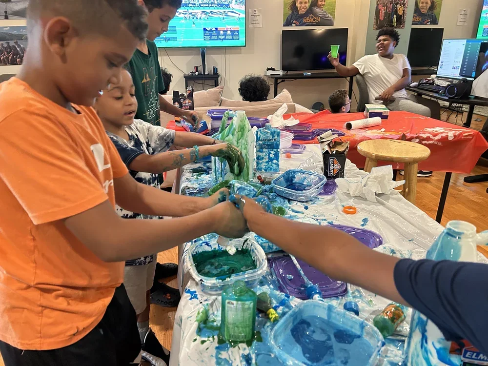 Children engaging in a sensory play activity with slime and water at a table, while other kids and a young woman watch and participate in the background.