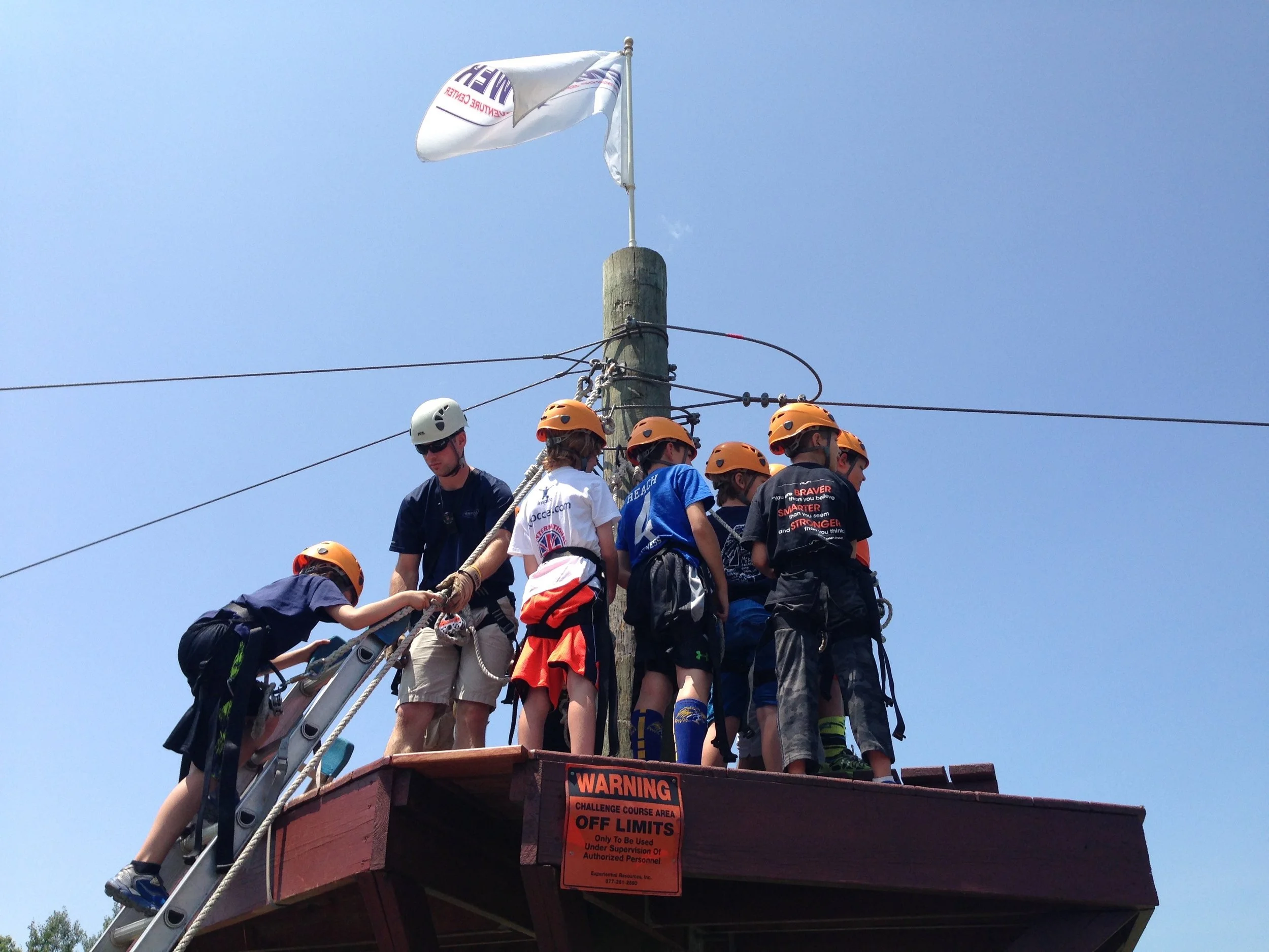 A group of kids and a supervisor wearing helmets standing on a challenge course platform, holding a rope, with a wooden pole and a flag flying in the background against a clear blue sky.