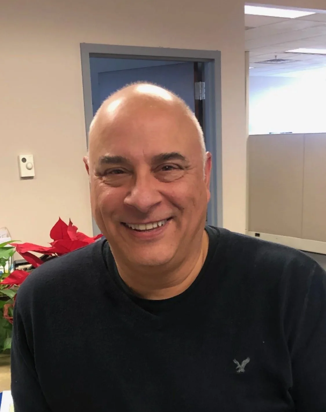Smiling man with a shaved head wearing a black sweatshirt, standing in an office with red poinsettia plants nearby.