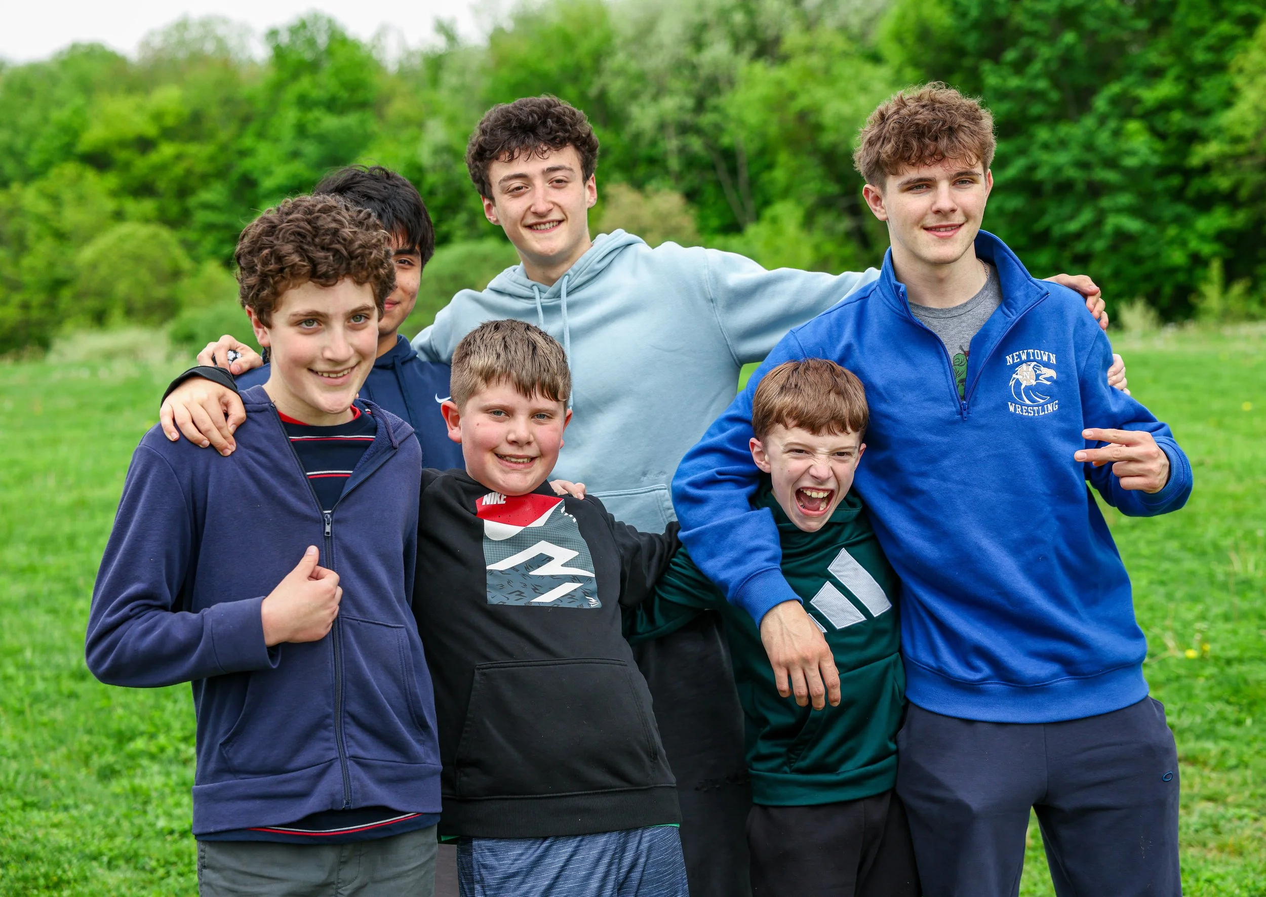 A group of seven boys standing outdoors on a grassy field with green trees in the background. They are smiling and appear happy, with some boys having their arms around each other.