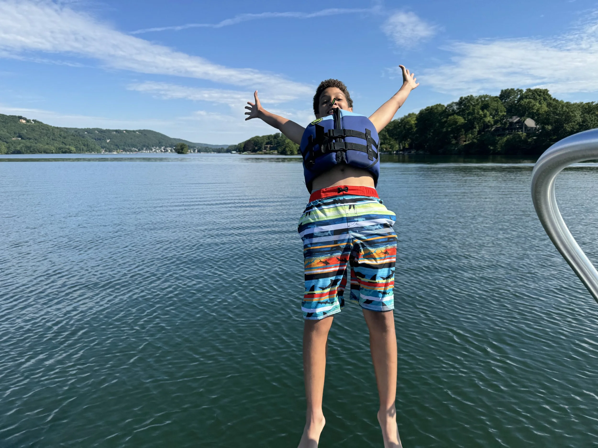 Young boy in colorful swim trunks and life jacket standing on a boat with outstretched arms, enjoying a sunny day on a calm lake surrounded by green hills and blue sky.