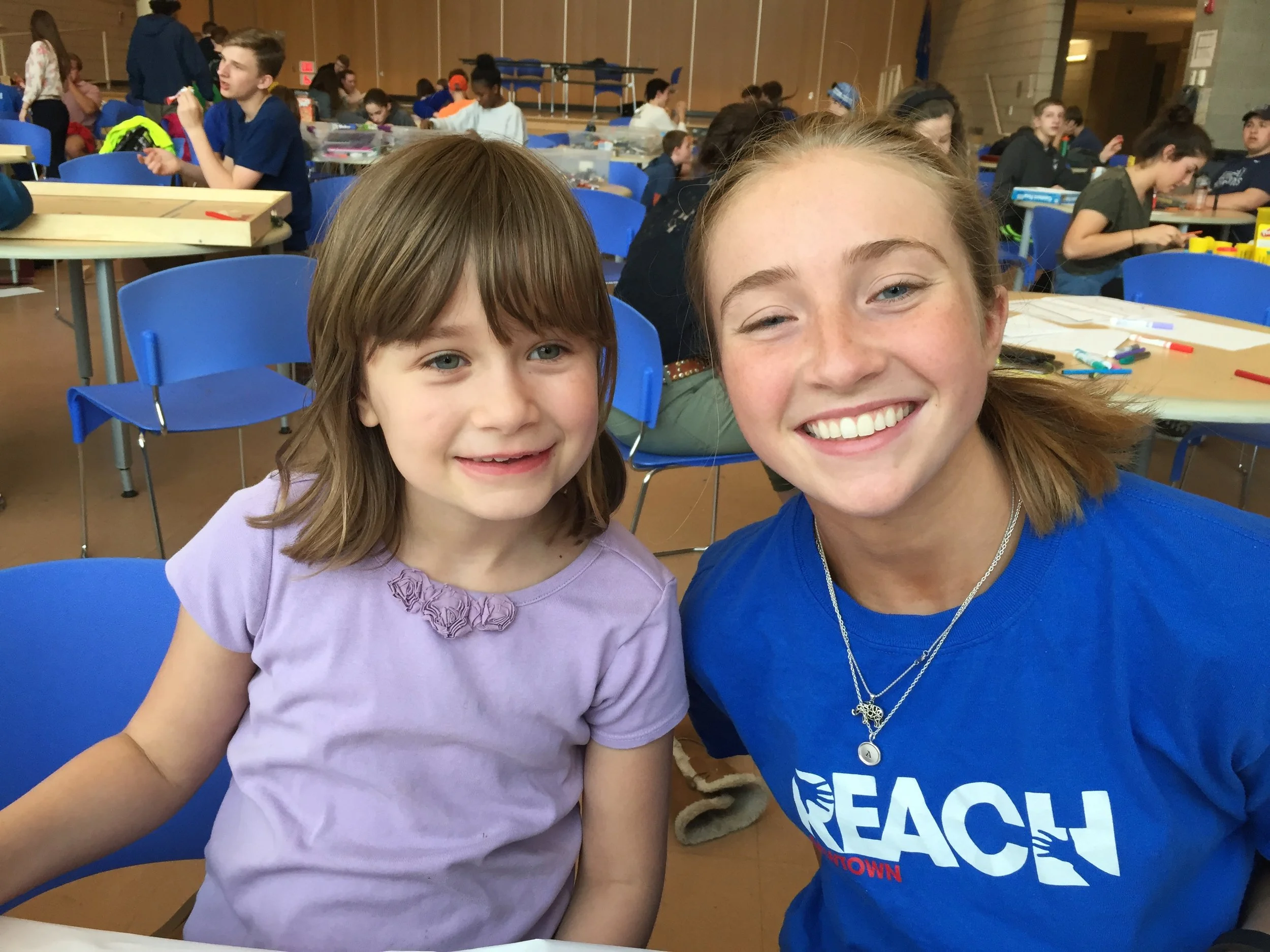 Two young girls smiling in a classroom or event setting, with one wearing a lavender shirt and the other wearing a blue EAOSH T-shirt. Several people and tables are visible in the background.