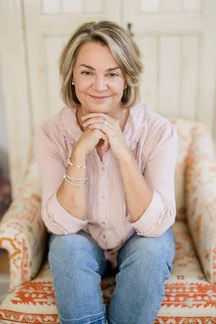 A woman with short blonde hair sitting on a patterned sofa with her hands clasped under her chin, smiling at the camera.