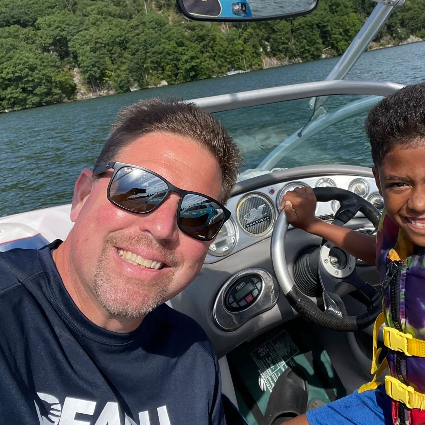 A smiling man with sunglasses taking a selfie on a boat with a young boy steering the boat, water and green trees in the background.