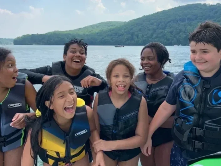 Six children in life jackets smiling and enjoying a boat ride on a lake with green, tree-covered hills in the background.
