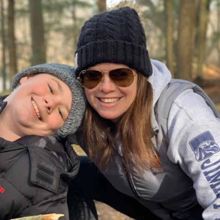 A woman and a child smiling and leaning their heads together outdoors in a wooded area, both wearing winter clothing including hats.