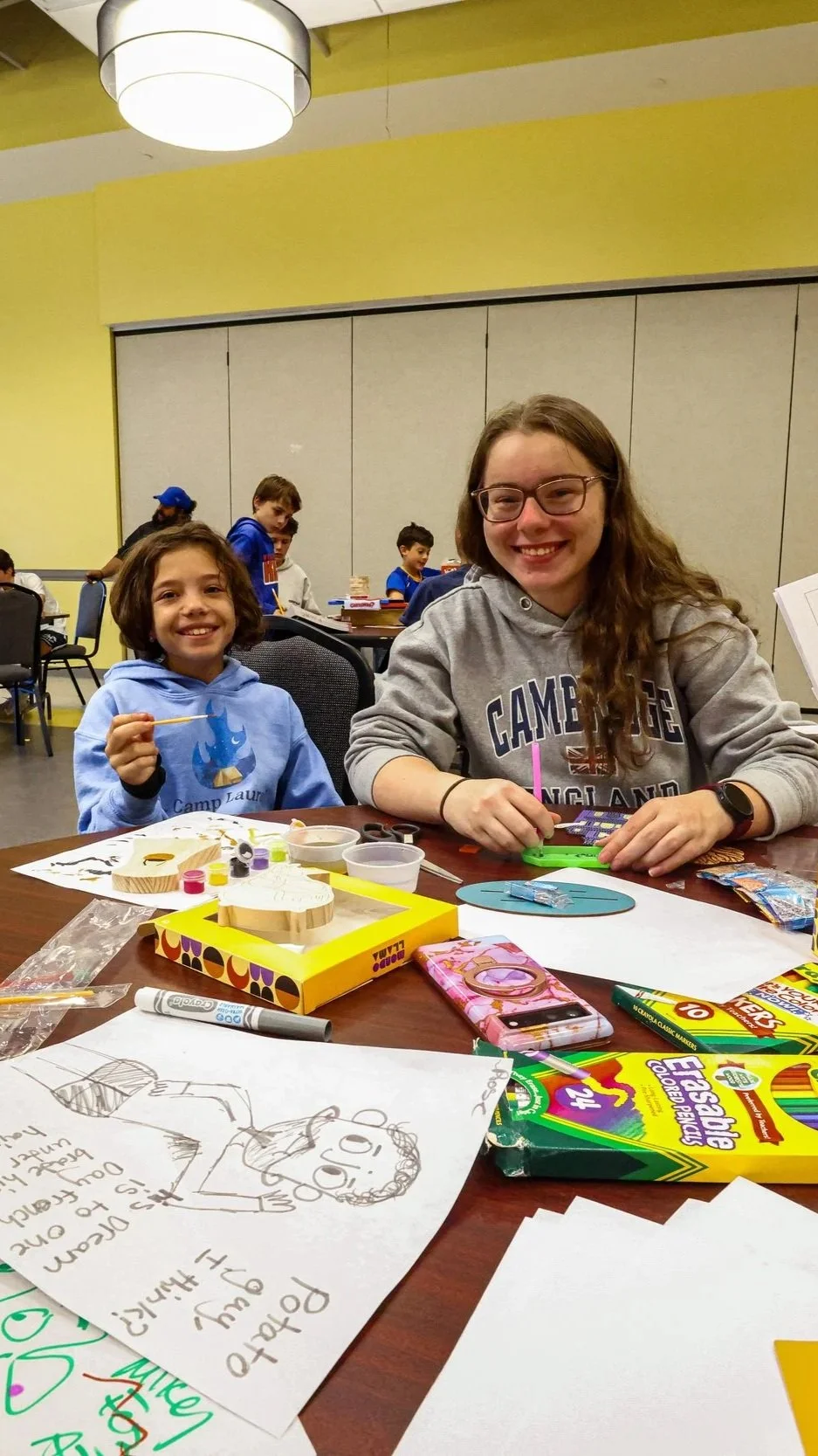 Two girls smiling at a table with craft supplies, coloring pages, and posters, in a room with a yellow wall and other children in the background.