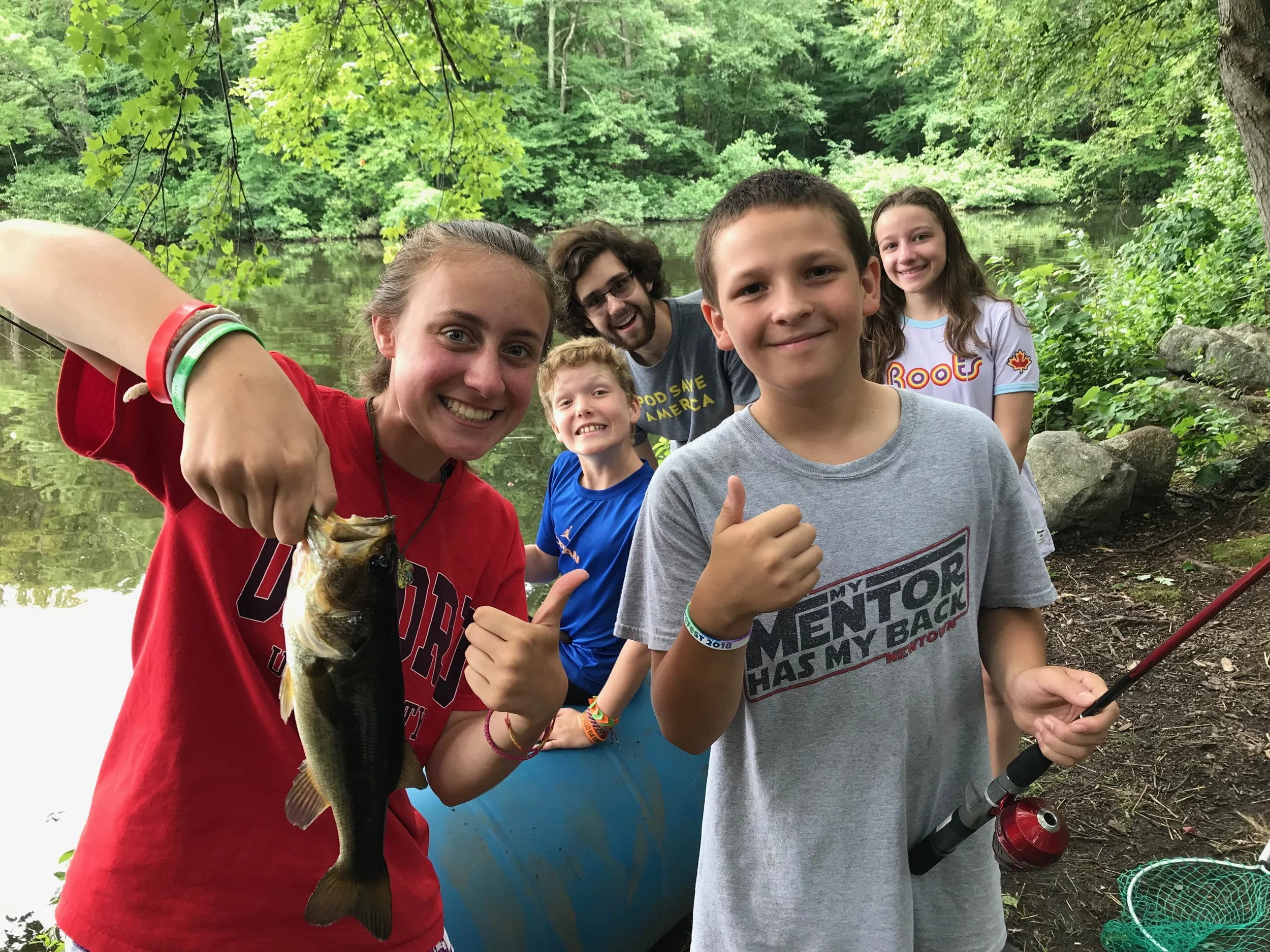 Group of five people near a body of water surrounded by trees, with one person holding a large fish.