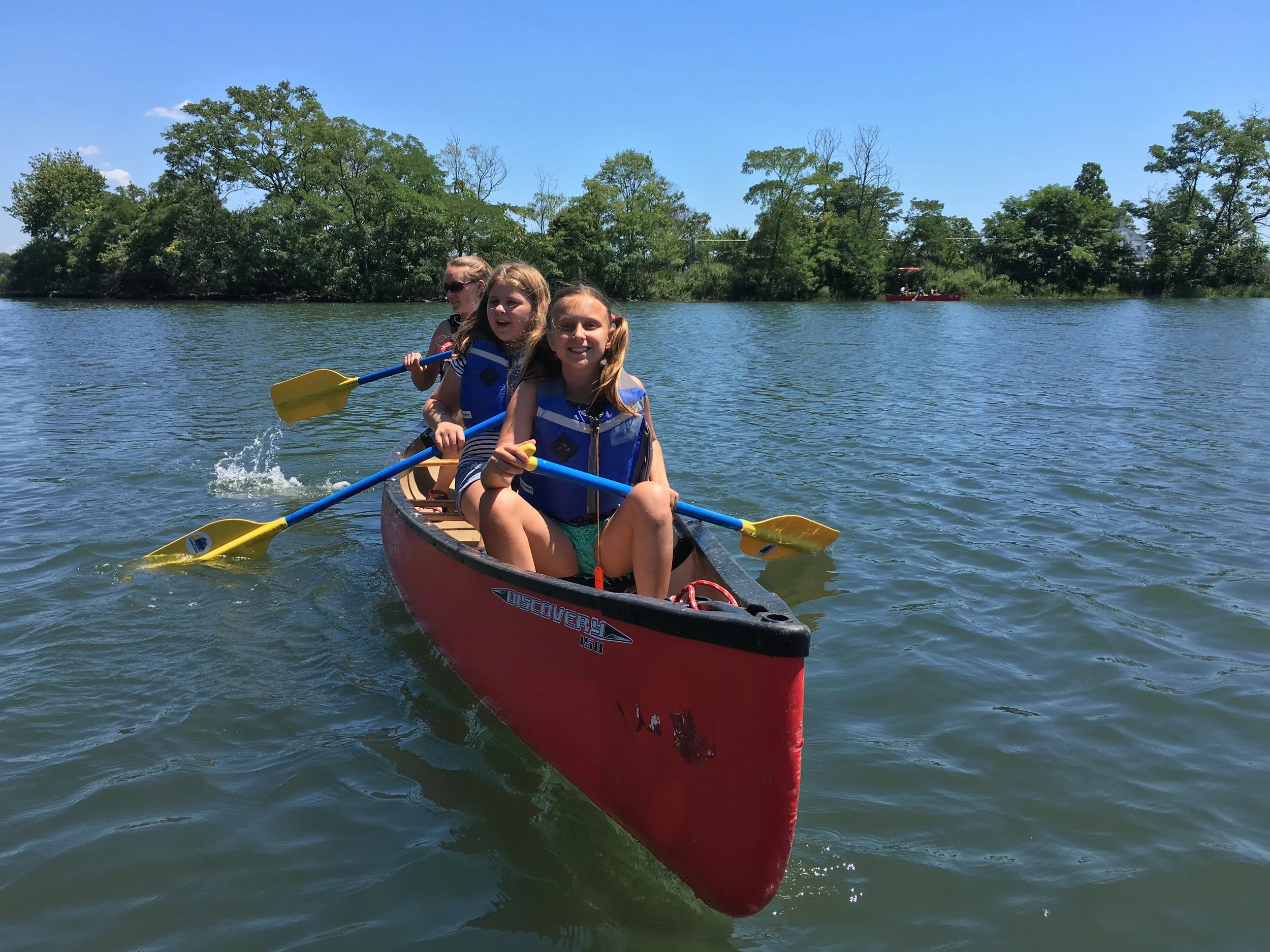 Three children wearing life jackets are paddling a red canoe on a lake. They are smiling and enjoying a sunny day with clear blue skies and trees in the background.