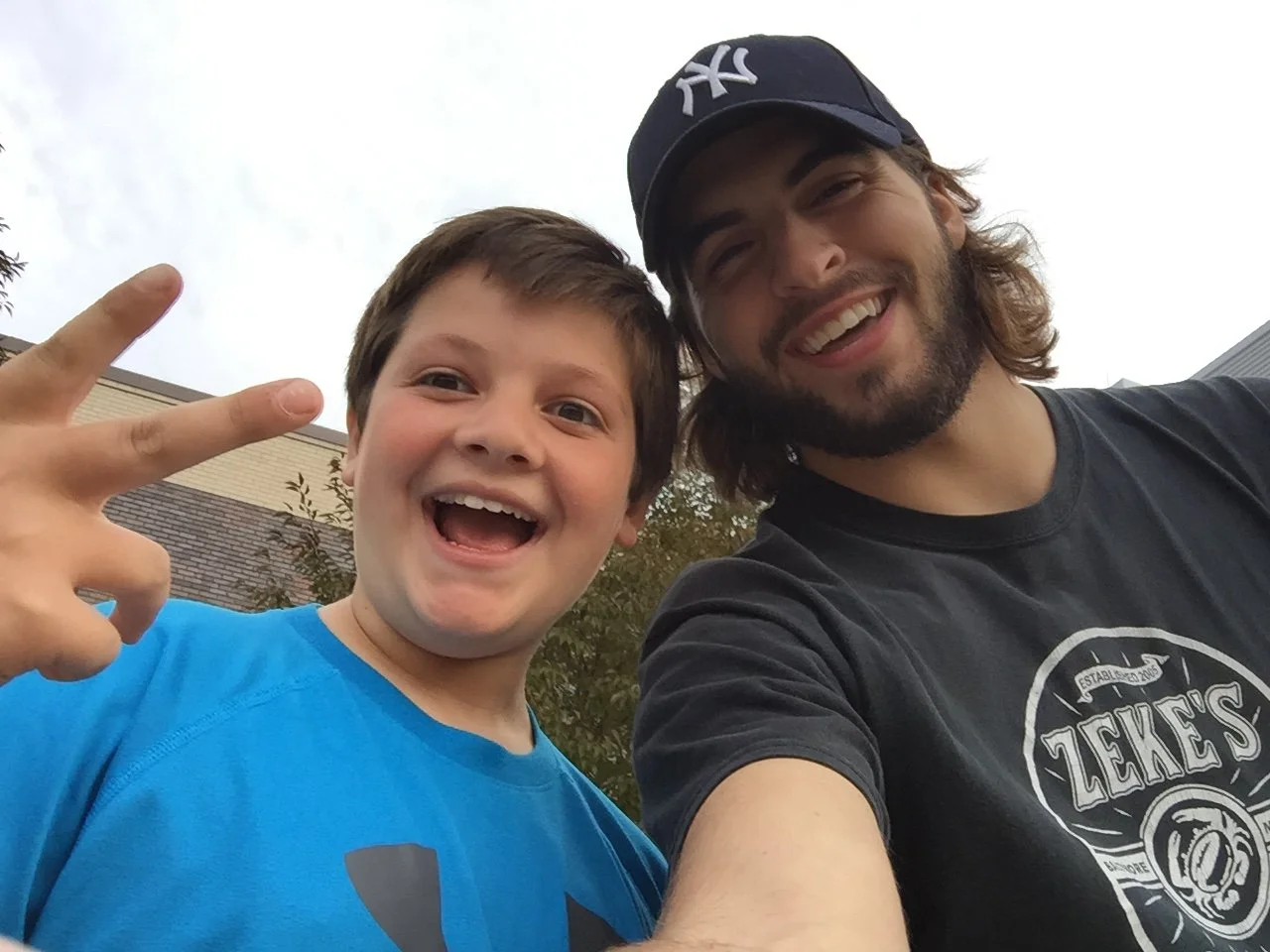 A young boy and an adult man taking a selfie together outdoors during daytime. The boy is making a peace sign with his fingers and smiling widely, wearing a blue shirt. The man has long hair, a beard, and is wearing a black baseball cap and black T-shirt with a brand logo. They appear happy and are standing in front of trees and a building.