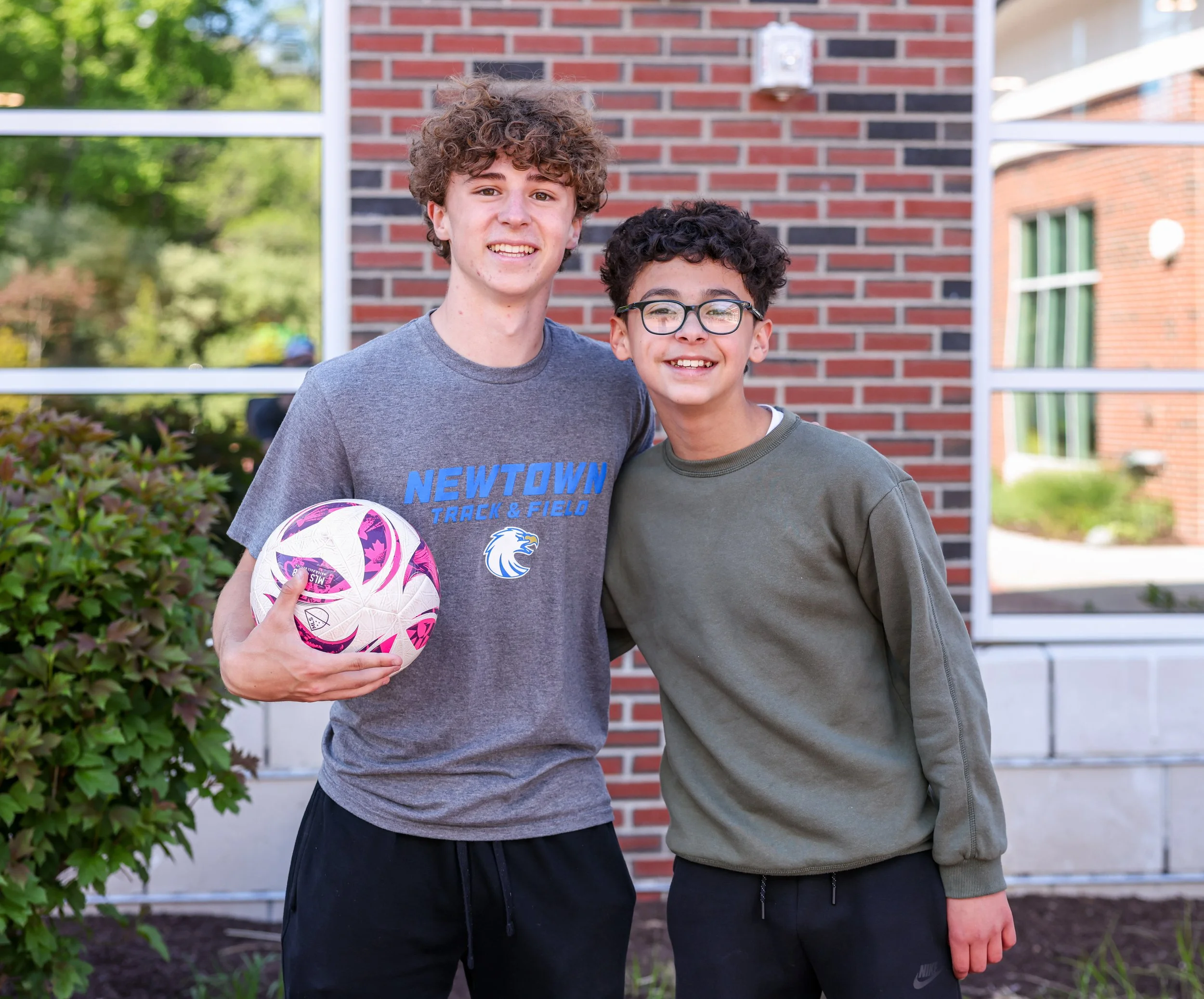Two boys standing outdoors, smiling at the camera. One holds a pink and white soccer ball. Behind them is a brick wall and greenery.