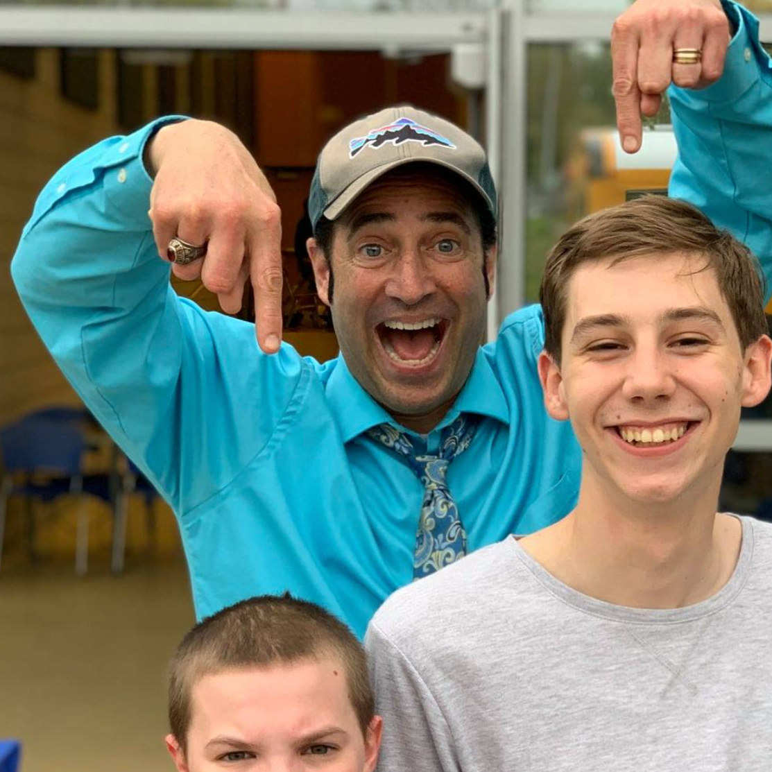 A man wearing a blue shirt, patterned tie, grey cap, and multiple rings is pointing at himself with both hands and has his mouth open in excitement. Two young boys are smiling in front of him, one with light brown hair and one with short dark hair. The background appears to be a casual indoor or outdoor setting.