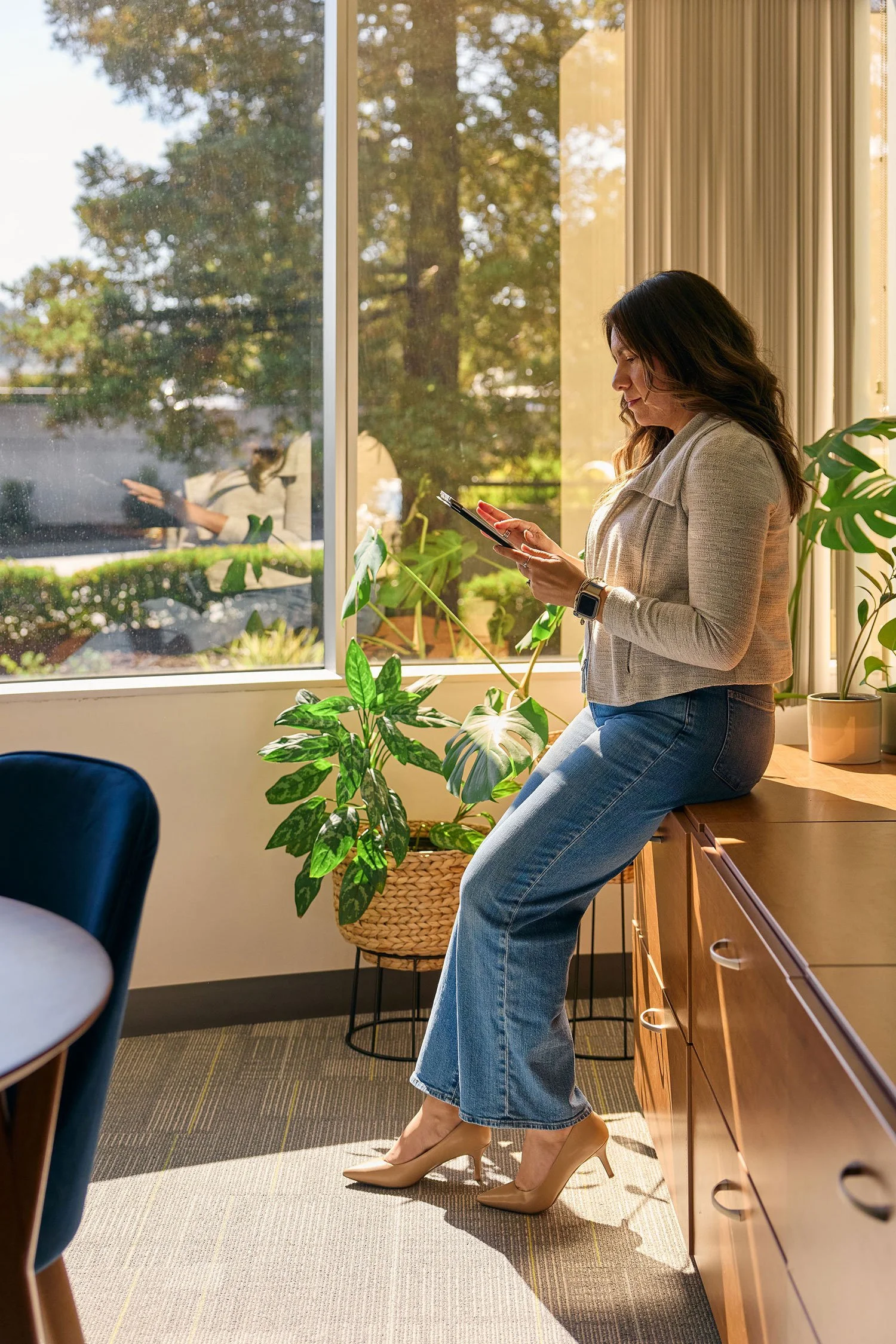A woman with long brown hair wearing a beige blazer, blue jeans, and beige high heels sitting on a wooden cabinet near a window looking at her smartphone inside a room with indoor plants.