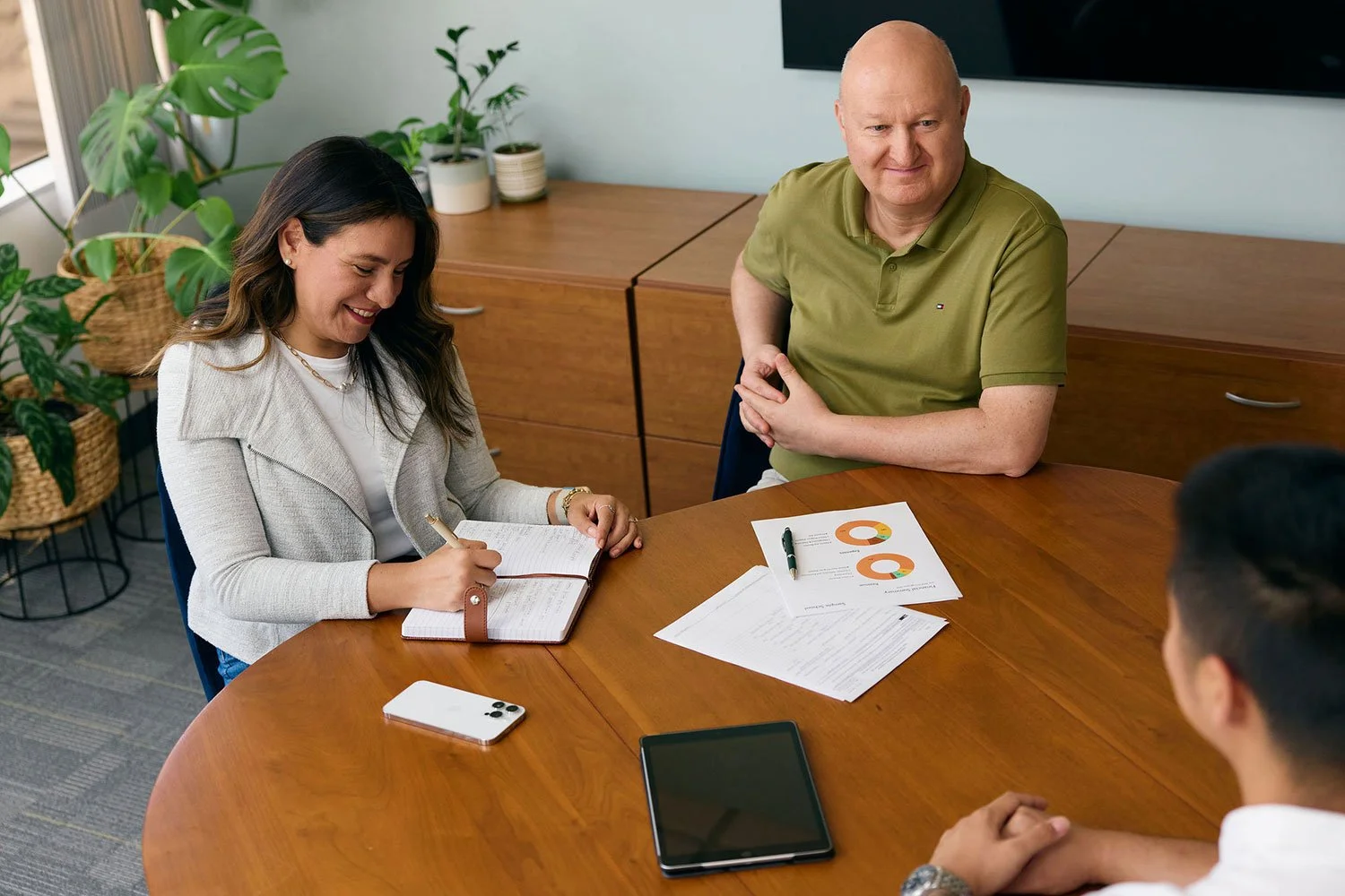 Four people sitting around a wooden table during a business meeting or discussion. The woman on the left is smiling, taking notes in a notebook, with a smartphone on the table. The man on the right is listening, with some documents and charts on the table. Two other people are partially visible in the foreground. The background includes green plants and a set of cabinets.