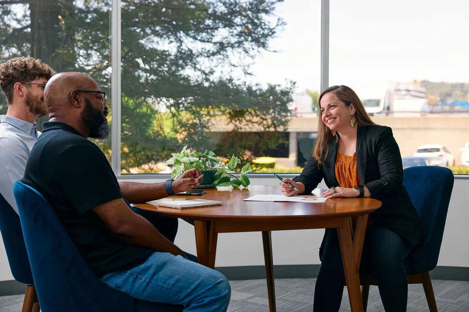 Three people sitting at a round wooden table in an office. A woman with long brown hair, black blazer, and orange top is smiling and holding a pen. Two men are sitting across from her; one with glasses and facial hair, the other bald with a beard. There is a potted plant, a notepad, and papers on the table, with a large window behind showing trees and parked cars outside.