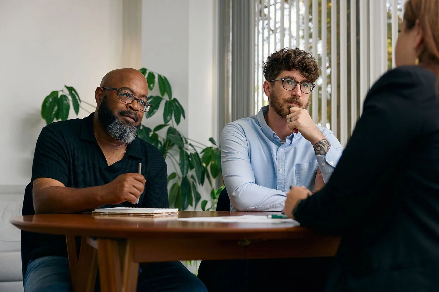 Three people sitting at a table during an interview or meeting, with a woman in a black blazer talking to two men, one with glasses and a beard, the other with glasses and curly hair, in a room with large windows and a plant in the background.