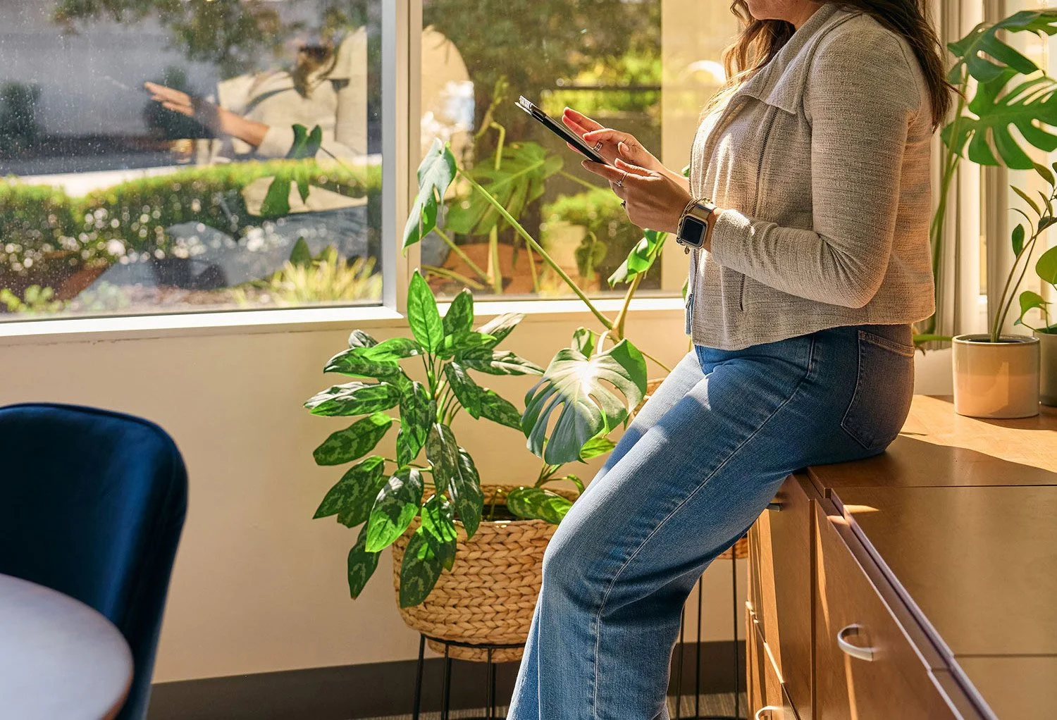 A woman leaning on a wooden cabinet, looking at her smartphone, inside a room with green houseplants and a large window showing an outdoor garden.