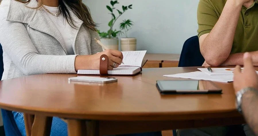 People sitting around a wooden conference table with notebooks, papers, and a tablet, in a room with potted plants.