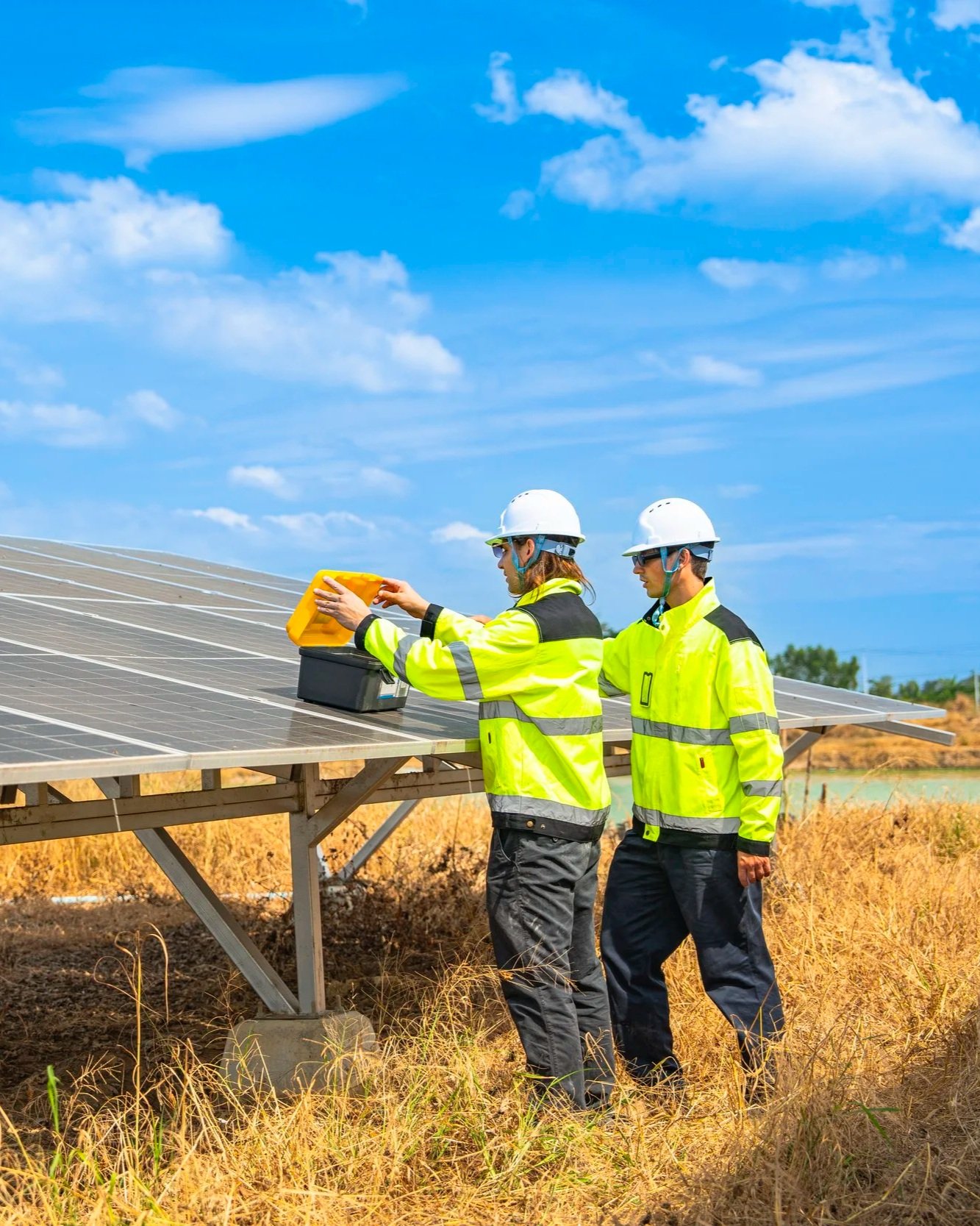 Two workers in high safety vests and helmets inspecting solar panels outdoors on a sunny day.