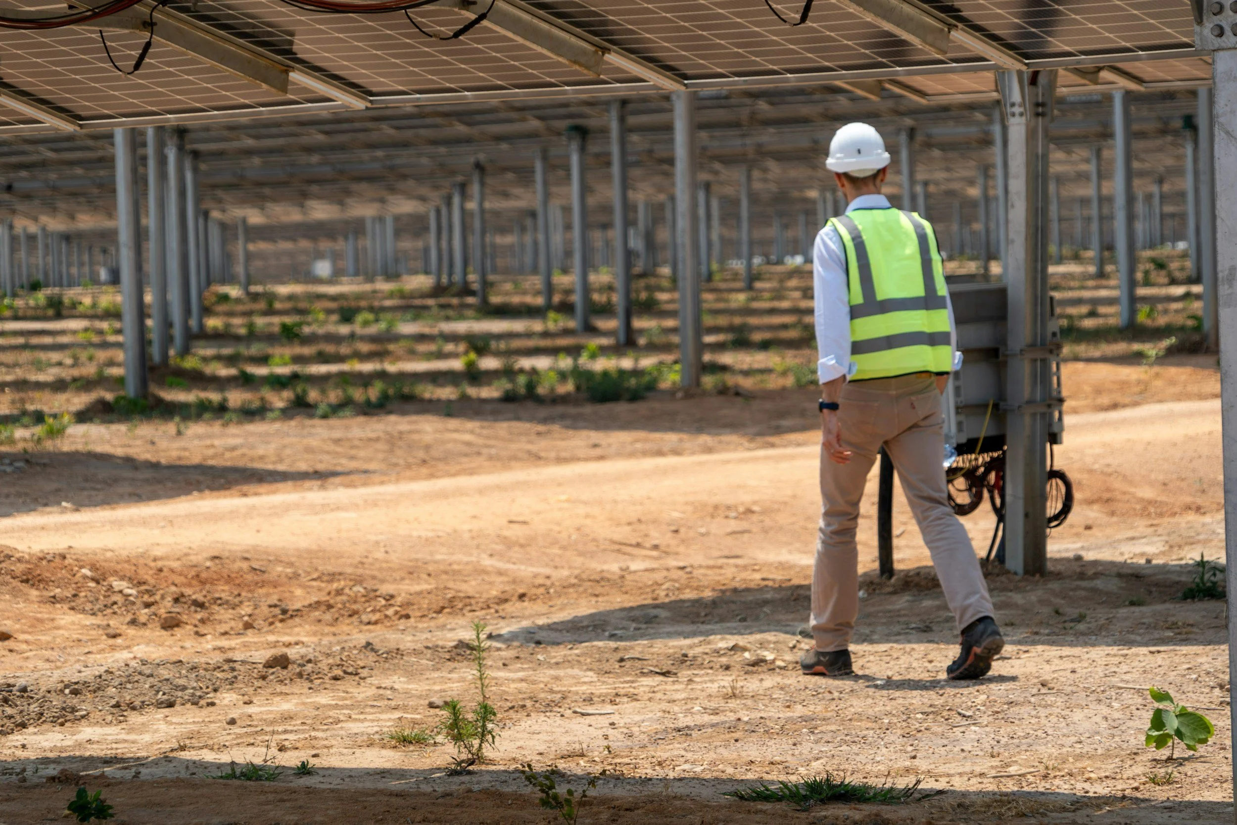A person wearing a white hard hat and a high-visibility safety vest inspecting a solar panel farm.