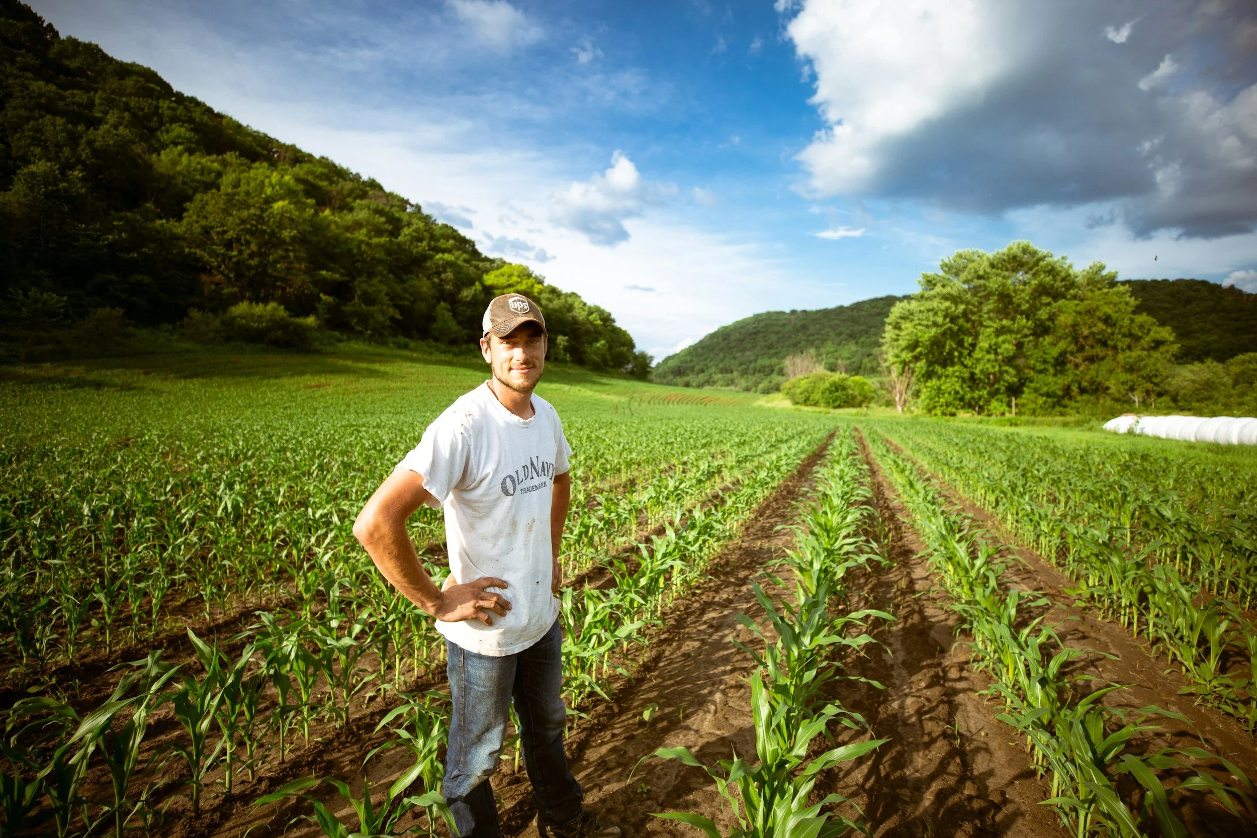 Man standing in a green farm field with crops, hills, trees, and a partly cloudy sky in the background.