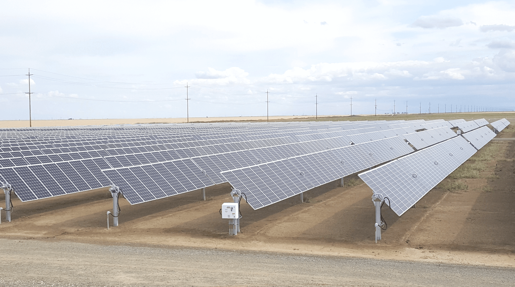 A large solar farm with multiple rows of solar panels installed on stands on a flat land, with power lines and poles in the background under a partly cloudy sky.