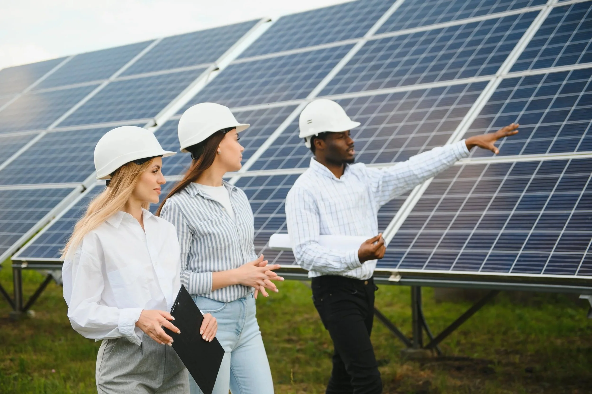 Three people wearing white safety helmets standing in front of solar panels, with one person explaining or pointing towards the solar panels.