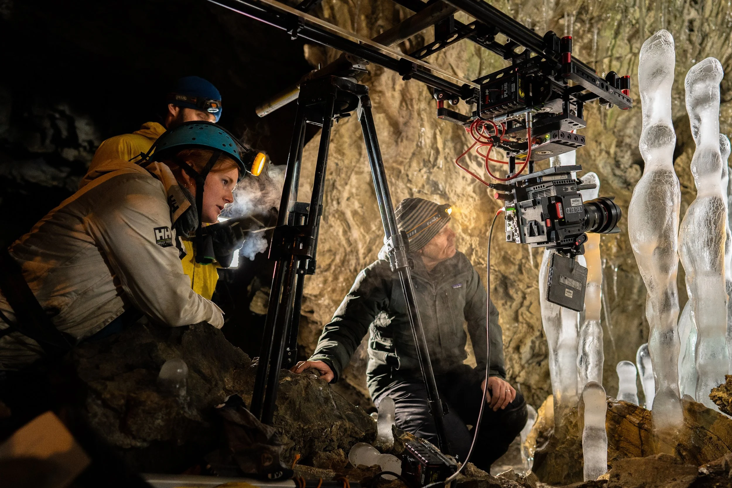 Filmmaking crew filming inside an ice cave, with ice stalagmites on the right and rocky walls, using a camera mounted on a rig and crew members dressed warmly.