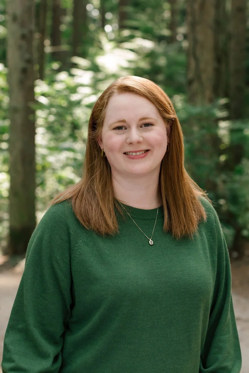 A woman with shoulder-length red hair smiling outdoors in a forest, wearing a green sweater and a silver necklace.