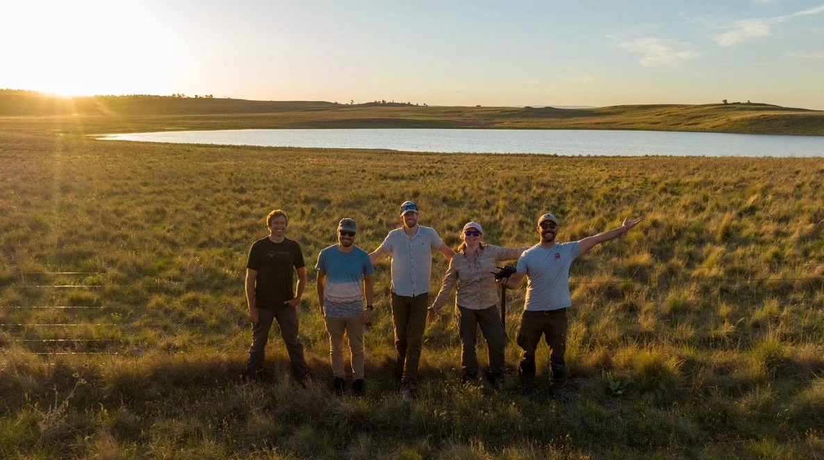 Five people standing in a grassy field near a small lake in Australia at sunset, smiling and posing for a photo.